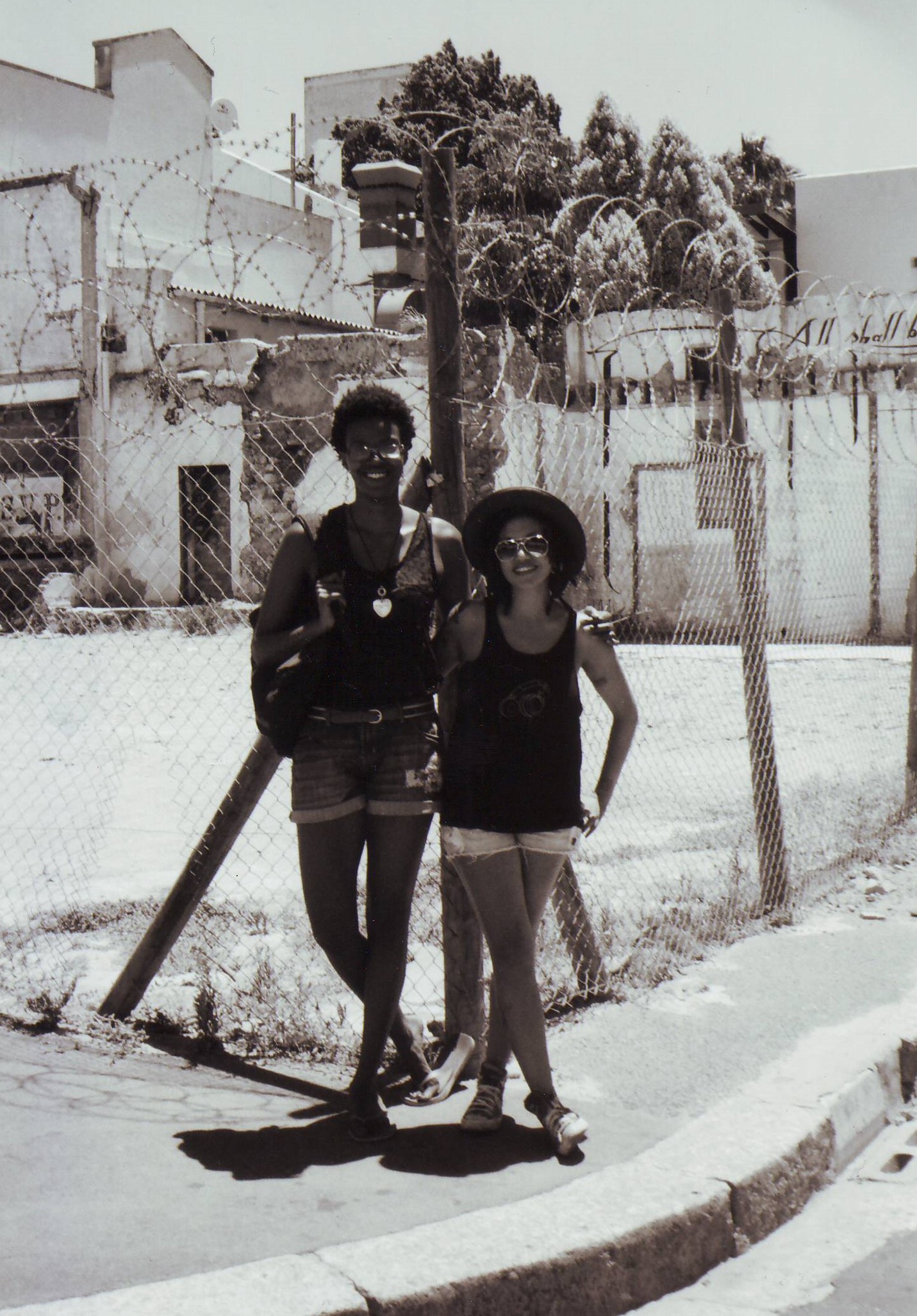 Zwei modisch gekleidete junge Frauen posieren für ein Foto. Im Hintergrund ein hoher Zaun mit Stacheldraht.