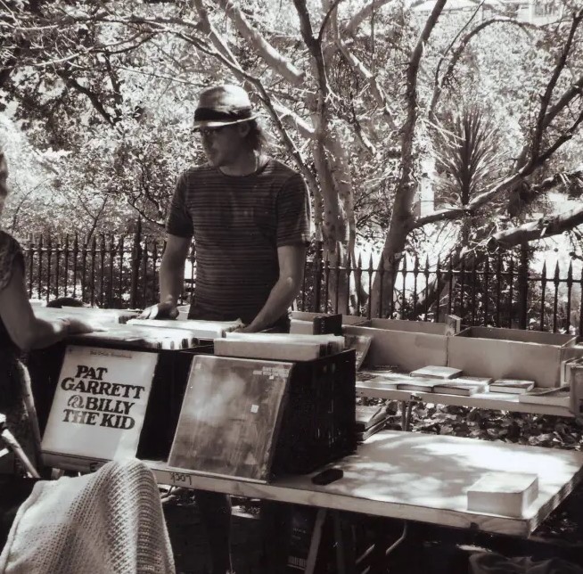 Ein Mann mit Hut hinter seinem Stand auf dem Flohmarkt. Er verkauft Schallplatten.